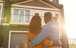 Young just-married couple standing at yard of house, hugging and with moving boxes in the background