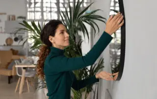 Close up smiling woman hanging or fixing mirror on wall, happy young female tenant renter decorating first own apartment, modern cozy living room, rent or mortgage concept, moving or relocation