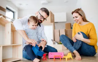 A portrait of young family with a toddler girl moving in new home, playing with moving boxes in the background.