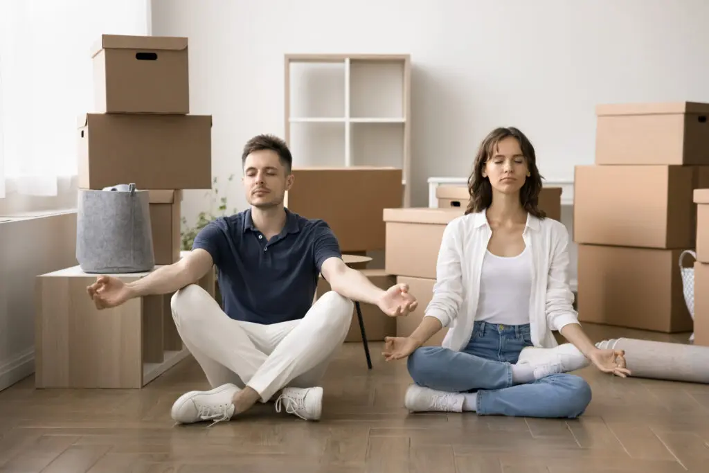 Young serene couple do meditation practice with eyes closed seated on floor on moving day, managing stress, relaxing, staying focused, maintain emotional balance on long, hard relocation, boxes nearby