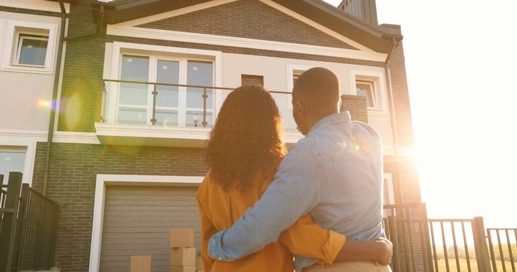 Young just-married couple standing at yard of house, hugging and with moving boxes in the background