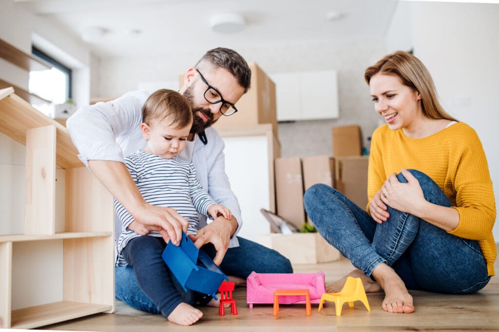 A portrait of young family with a toddler girl moving in new home, playing with moving boxes in the background.