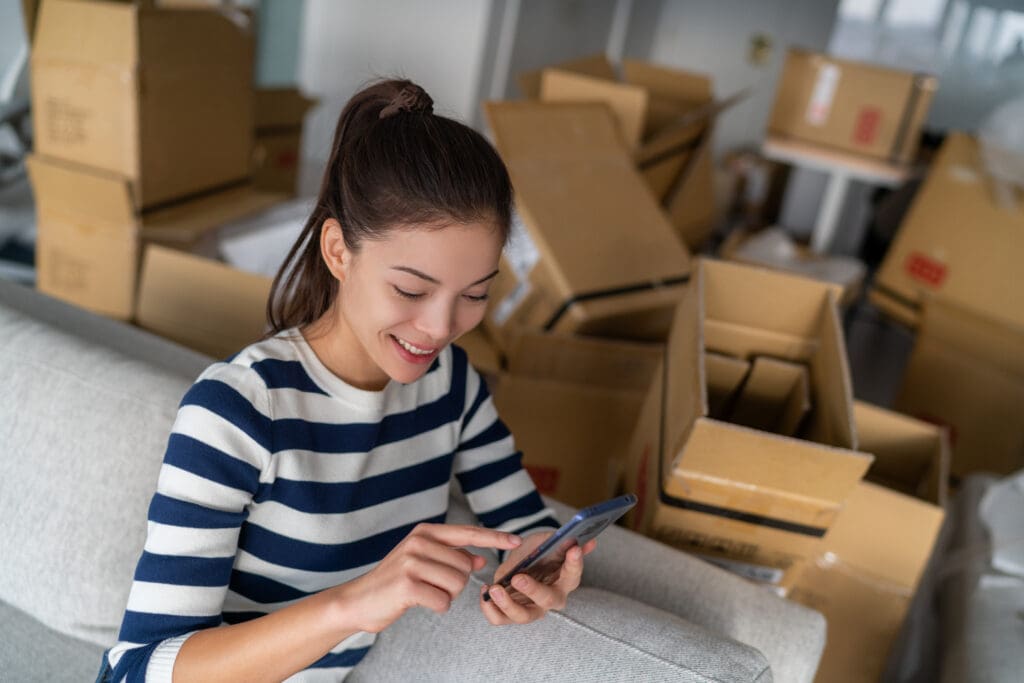 woman using cellphone mobile phone for to plan move with boxes around her