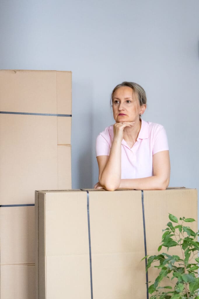 Woman leaning on moving boxes while thinking about future life changes. Relocation, emotional transition, new beginnings and uncertainty after moving to new home.