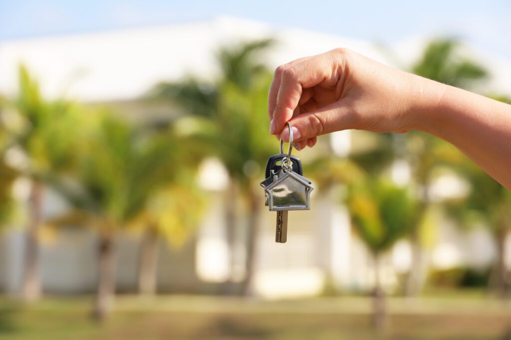 home keys in female hand on background of house surrounded by palm trees. Buying or rental a home on ocean coast, removal