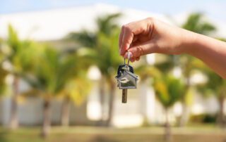 home keys in female hand on background of house surrounded by palm trees. Buying or rental a home on ocean coast, removal